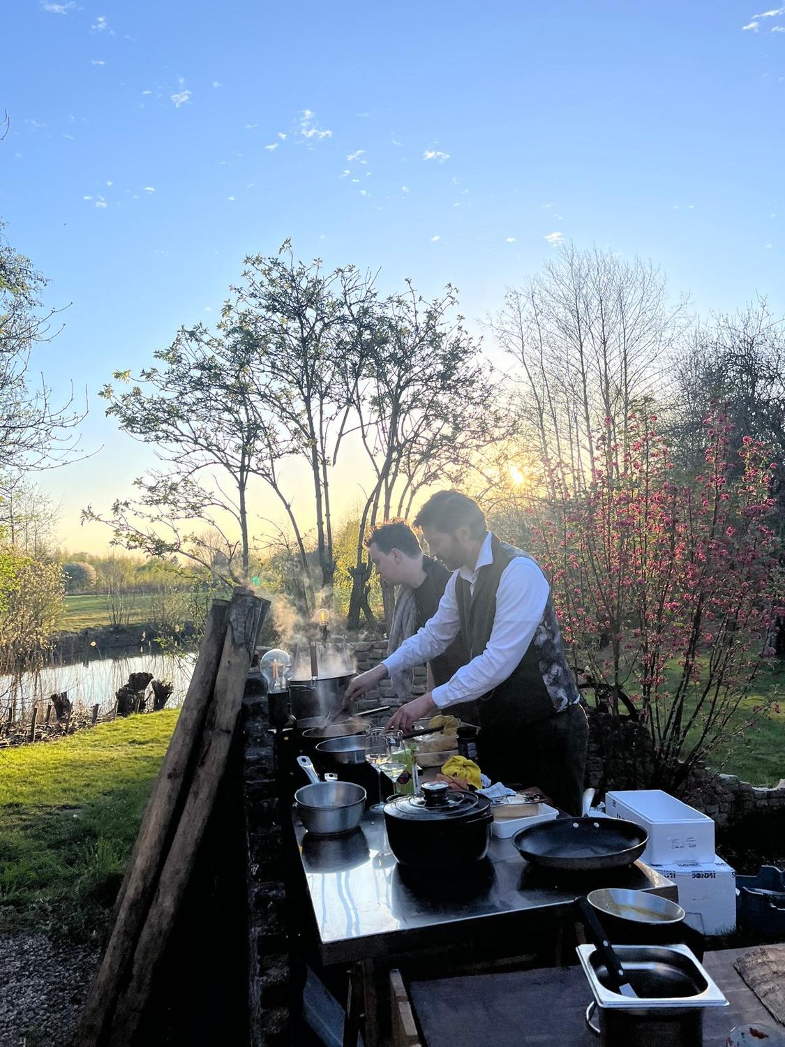 Twee koks aan het werk in de buitenkeuken bij zonsondergang, naast de vijver
